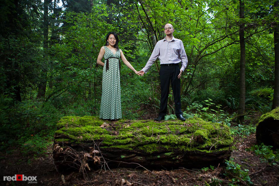 Nobuyo and Rory  stand on a large, moss-covered log during their engagement portrait session at Seward Park in Seattle. (Photography by Andy Rogers/Red Box Pictures)