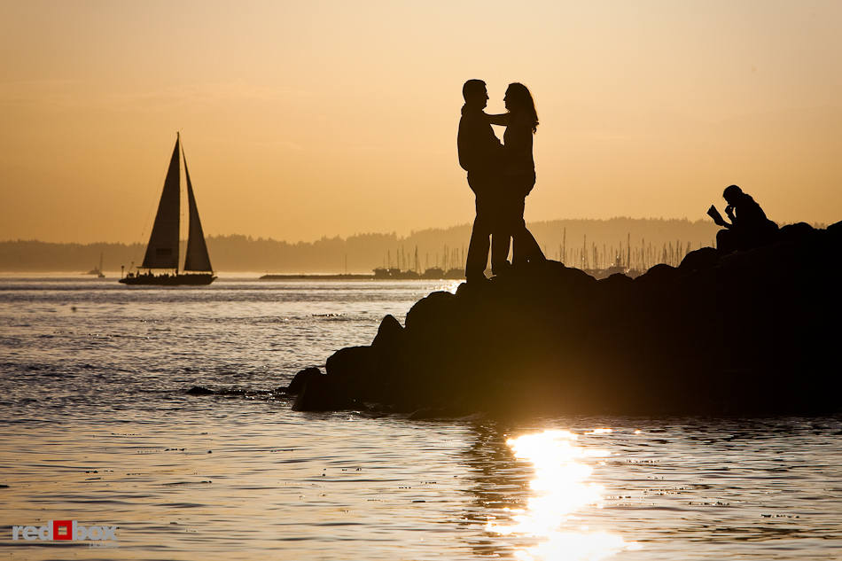 Katherine and Bryan hug while the sun sets and sailboats glide over Elliott Bay during their engagement session at Myrtle Edwards Park in Seattle. (Photo by Dan DeLong/Red Box Pictures)