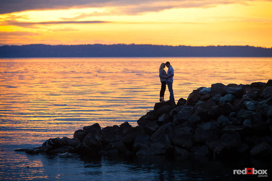 With a striking sunset over the water of Puget Sound behind them, Tyler and Megan hug on the waterfront in Edmonds, WA during their engagement photography session. (Photo by Dan DeLong/Red Box Pictures)